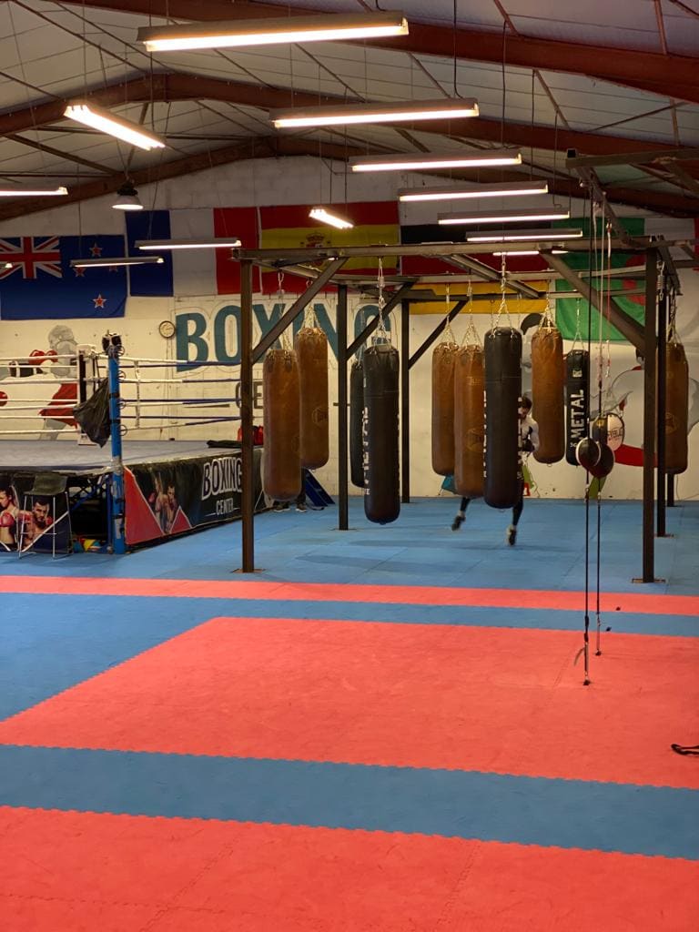 La salle vue en longueur : les drapeaux au plafond, le mot « Boxing » peint au mur, le ring à gauche et le portique de sacs au centre.